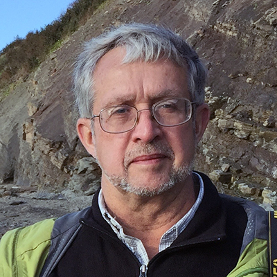A man with short grey hair wearing a windbreaker and square glasses stands in front of a rocky cliff outside.